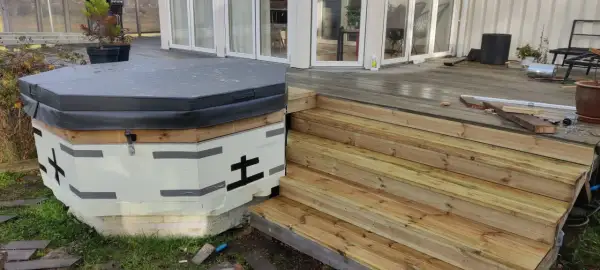 Pågår bygga badtunna. Isolerad stomme med trädetaljer och en grå presenning. Nya trätrappor leder upp till altanen.