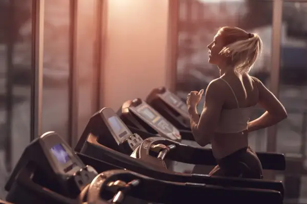 Frau mit blondem Pferdeschwanz walkt auf dem Laufband im Fitnessstudio, das warme Abendlicht fällt durch die Fenster.