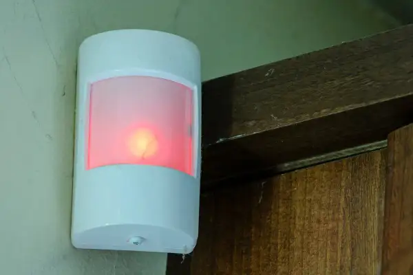 Biały czujnik ruchu z czerwoną diodą, zamontowany na ścianie obok drewnianej framugi. Pokazuje, jak działa czujnik ruchu.