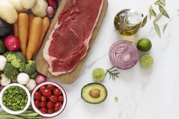 Un corte de carne magra, rodeado de verduras frescas como zanahorias, brócoli, tomates y aguacate, junto a aceite de oliva y limón.