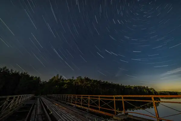 Nocne niebo nad starym mostem, gdzie gwiazdy kreślą na ciemnym tle świetliste ślady.