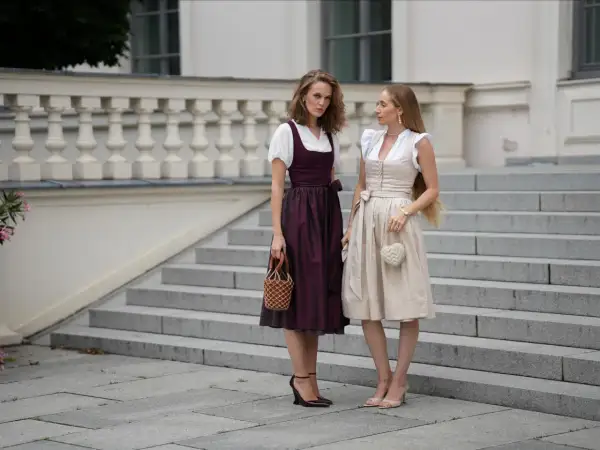 Zwei Frauen in Dirndln vor einer Treppe. Die eine trägt einen dunkelroten Dirndl mit Spitzenbesatz am Rock, die andere einen hellbeigen. Dirndl unterrock ja oder nein? Hier wird die Frage stilvoll beantwortet.