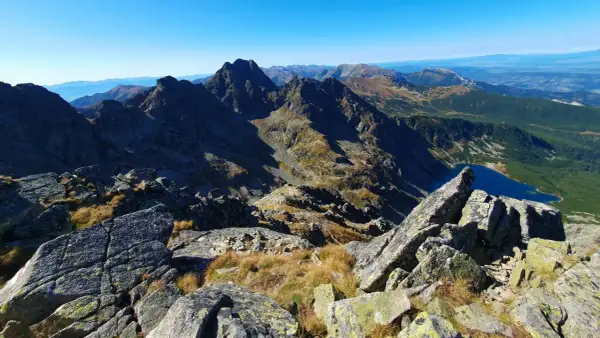 Widok z kamienistego szczytu w Tatrach Wysokich na skaliste granie, trawiaste zbocza i turkusowe jezioro.