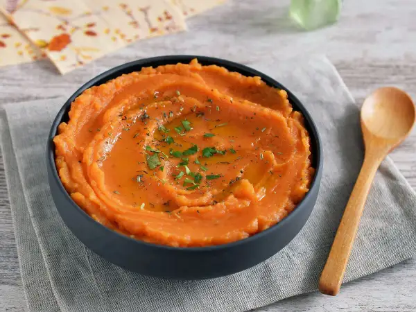 Un tazón de puré de boniato gourmet, cremoso y adornado con aceite y hierbas. Una cuchara de madera descansa a su lado.