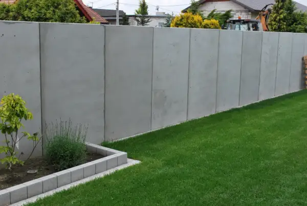 Nowy, wysoki betonowy płot dzieli działki, symbolizując konflikt z sąsiadem o ogrodzenie.