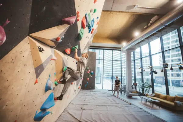 Une femme grimpe sur un mur de bloc dans un centre d'escalade moderne, le climbing district Batignolles. Un homme observe depuis le sol.