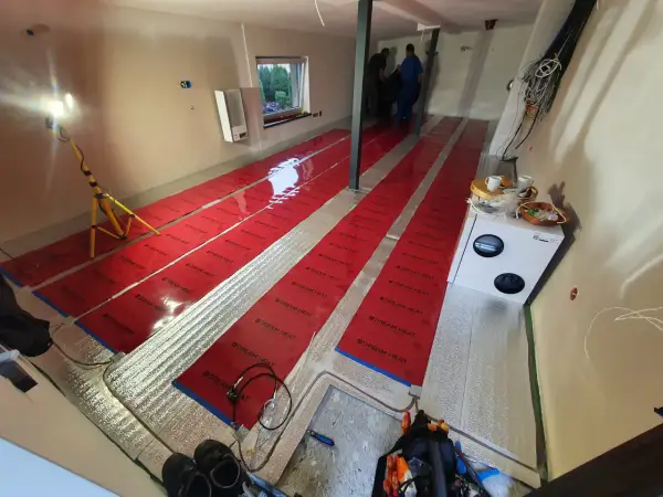 Instalacja folii grzewczej. Widać długie pasy czerwonej folii na podłodze, przygotowane do montażu. Wady tej metody mogą obejmować nierównomierne rozprowadzanie ciepła.