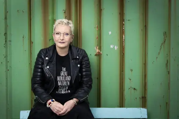 Ola Liljedahl, med kort blont hår och glasögon, sitter framför en sliten grön vägg. Hon bär en svart läderjacka och en t-shirt med texten "THE GREAT NORRLAND NATUREN".