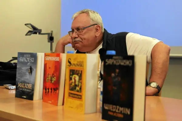 Mężczyzna zamyślony nad stosem książek, wśród nich "Wiedźmin" i inne tytuły Sapkowskiego, idealne dla fanów szukających książek podobnych do Wiedźmina.