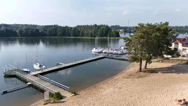 Wdzydze Kiszewskie plaża z pomostami i żaglówkami. Piasek, woda i zielone drzewa tworzą idylliczny krajobraz.