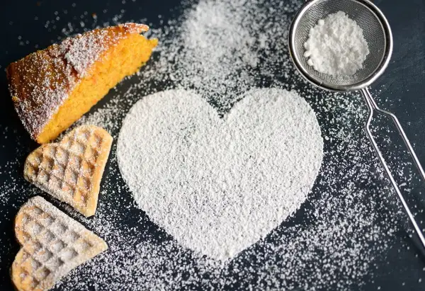 A slice of cake, heart-shaped cookies, and a heart shape made of powdered sugar on a dark surface.