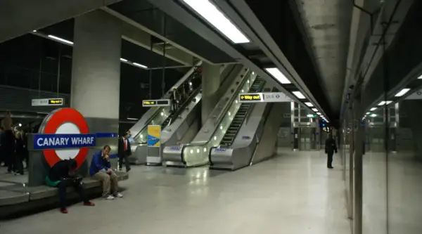 Stacja Canary Wharf w **metro londyn** z charakterystycznym znakiem, ruchomymi schodami i ludźmi czekającymi na pociąg.