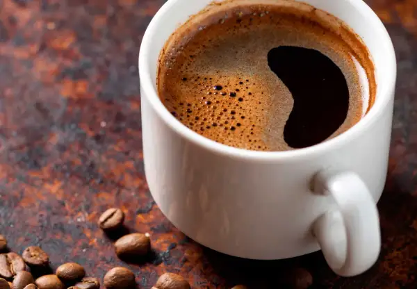 En vit kopp med mörkt kaffe och skum. Kaffebönor ligger utspridda. Undrar du hur mycket koffein i en kopp kaffe?
