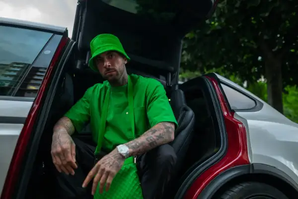 Malik Montana w zielonym kapeluszu i koszulce, siedzi w samochodzie z otwartymi drzwiami. Wygląda jak adwokat diabła, gotowy na nowe wyzwania.