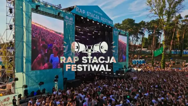 Tłum ludzi na koncercie rap festiwal. Na scenie wielkie ekrany z napisem "RAP STACJA FESTIWAL" i grafiką megafonów.