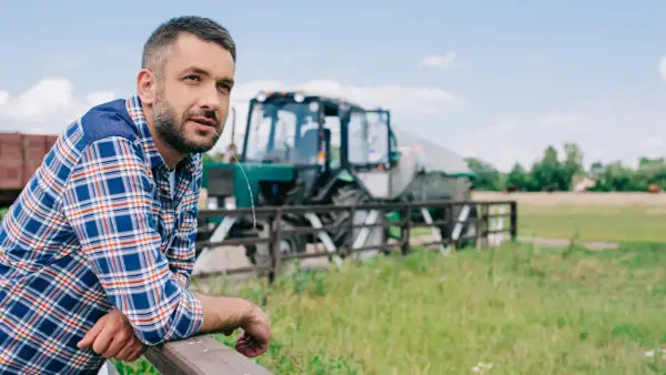 Rolnik w kratę oparty o płot, w tle traktor. Wsparcie dla małych gospodarstw to klucz do rozwoju wsi.
