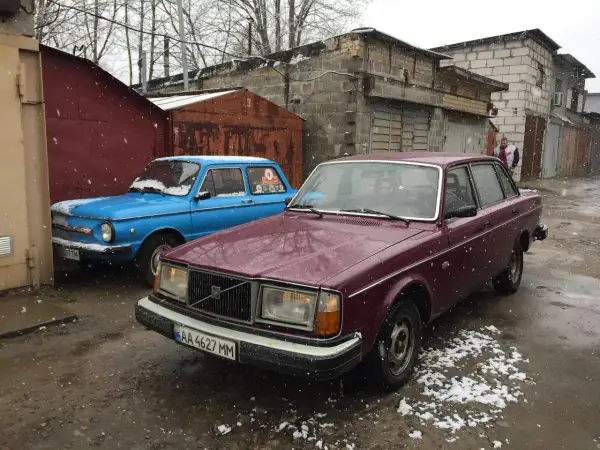 En vinröd Volvo 244 DLS står parkerad bredvid en blå bil i snöfall.