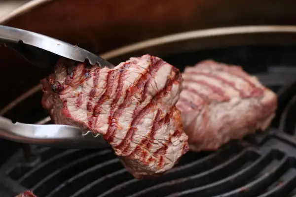 Zangen halten ein saftiges Stück Roastbeef über einem Grillrost. Das Fleisch hat schöne Grillstreifen und ist perfekt gegrillt.