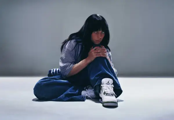Billie Eilish siedzi na podłodze w luźnych dżinsach i koszuli, patrząc w obiektyw.