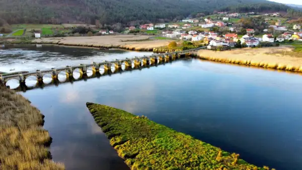 Un puente de piedra con arcos cruza un río azul, conectando un pueblo pintoresco con campos verdes. El puente, conocido como Ponte nafonso, se alza majestuoso sobre el agua.