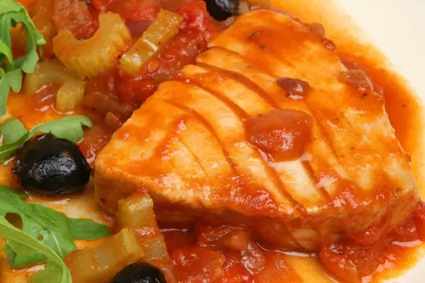Un succulento tonno in padella con pomodoro, olive nere e sedano, guarnito con rucola fresca.