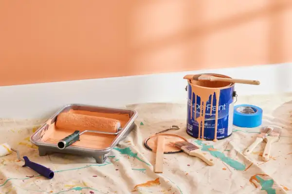 Painting supplies, including a roller in a tray and a can of peach-colored satin paint, are laid out on a drop cloth.