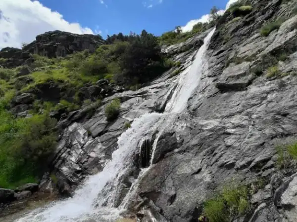 Una hermosa cascada en Madrid, cayendo por rocas grises y vegetación verde bajo un cielo azul con nubes blancas.