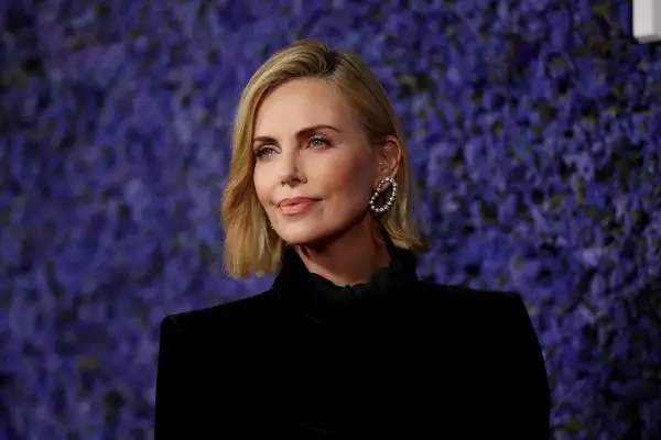 Charlize Theron, bekannt für ihre Rollen in Filmen wie "Mad Max: Fury Road", posiert vor einem lila Hintergrund.