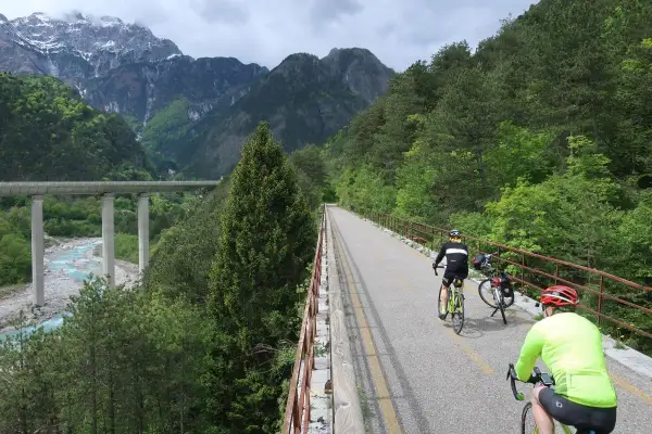 Rowerzyści przemierzają malowniczą trasę **Alpe Adria** w otoczeniu gór i zielonych lasów. Widoczny most i rzeka dodają krajobrazowi majestatu.