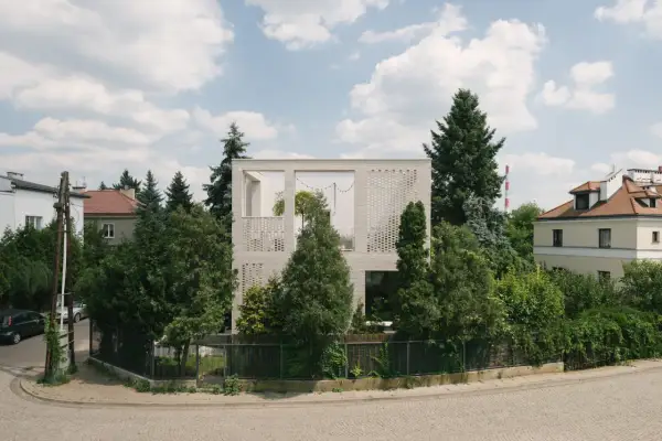 Nowoczesny dom typu kostka z białej cegły, otoczony zielenią. Widać fragmenty sąsiednich budynków i błękitne niebo z chmurami.
