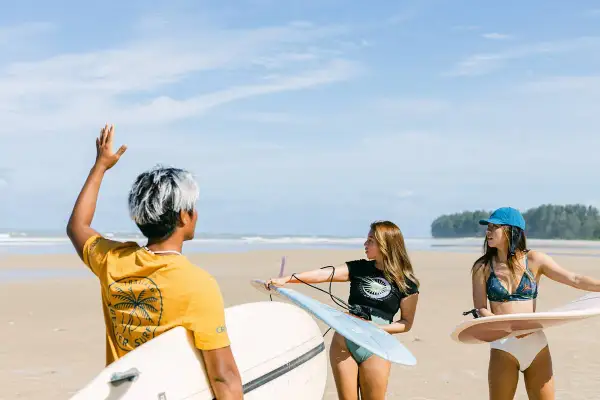 Tre vänner med surfbrädor på stranden i Thailand. En man med vitfärgat hår vinkar, medan två kvinnor förbereder sig för att surfa.