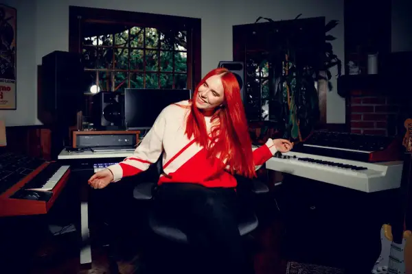 Noonie Bao, with vibrant red hair, smiles and gestures in a music studio filled with synthesizers and speakers.