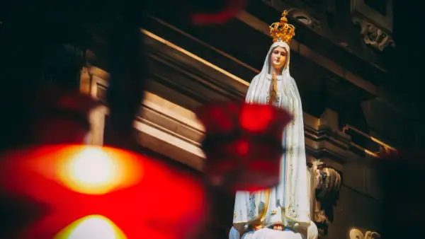 Figurka Matki Bożej Fatimskiej w koronie, z rękami złożonymi do modlitwy. W tle widać architekturę kościoła, a na pierwszym planie rozmyte czerwone światła, symbolizujące trzecia tajemnica fatimska.