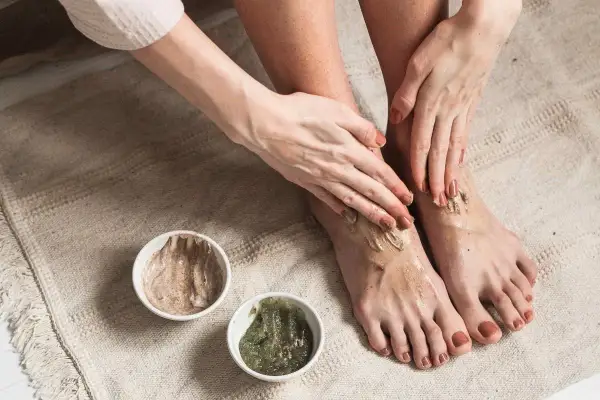 Kobieta aplikuje na stopy peeling, przygotowując je do użycia skarpetek złuszczających. Dwie miseczki z kosmetykami stoją obok.