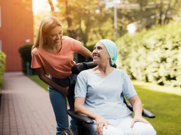 Uśmiechnięta kobieta w chuście na głowie siedzi na wózku inwalidzkim, a młoda kobieta pcha go. Nadzieja, że czy rak 4 stopnia jest wyleczalny, dodaje im sił.