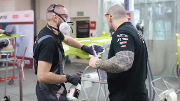 Dwóch mężczyzn w maskach i rękawiczkach pracuje nad zielonym elementem, sprawdzając, czy na podkład epoksydowy można kłaść lakier.