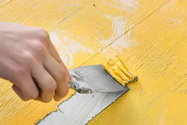 Mano raspando pintura amarilla vieja con una espátula. El decapado de pintura revela madera debajo.