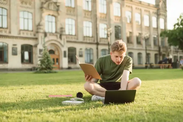 Joven estudiando en el césped frente a un edificio histórico, con un libro y portátil. Un momento de aprendizaje y **intercambio cultural** en un entorno inspirador.