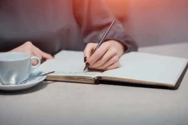 Dłoń pisze w notesie, szukając inspiracji, jak napisać intencję za zmarłego wzór. Obok filiżanka kawy.