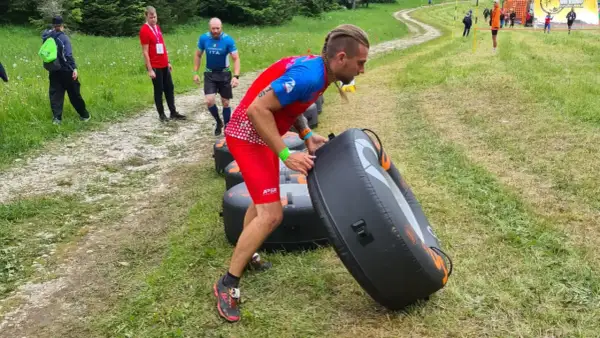 Uomo con treccia si allena al parco, spingendo un grande gonfiabile nero. Altri partecipanti corrono su un sentiero erboso.