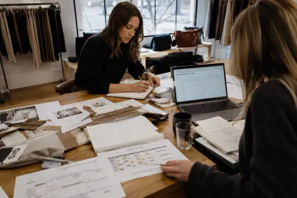 Dos mujeres revisan proyecciones de ventas y calendarios de producción. Una escribe en una libreta, la otra señala un documento. Se preguntan qué significa sin stock.