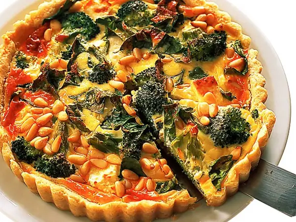 En gyllene vegetarisk paj med broccoli, spenat och pinjenötter. En bit lyfts upp med en pajspade.