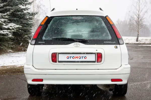 Biała Toyota Corolla E11 kombi z napisem "OTOMOTO" na tablicy rejestracyjnej, stoi na mokrym asfalcie podczas opadów śniegu.