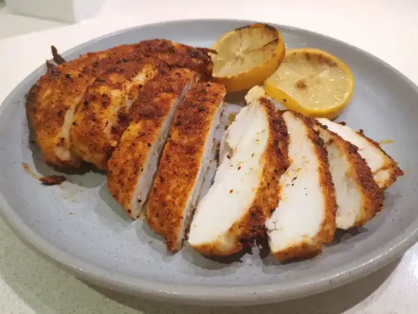Skivad panerad kyckling med kryddig yta, serverad med grillade citronklyftor på en grå tallrik.