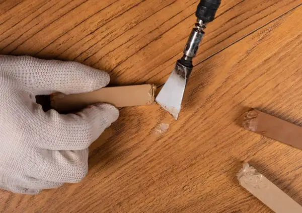 Mano guantata rimuove schegge da un parquet rovinato, mostrando rimedi fai da te per riparare il legno danneggiato.
