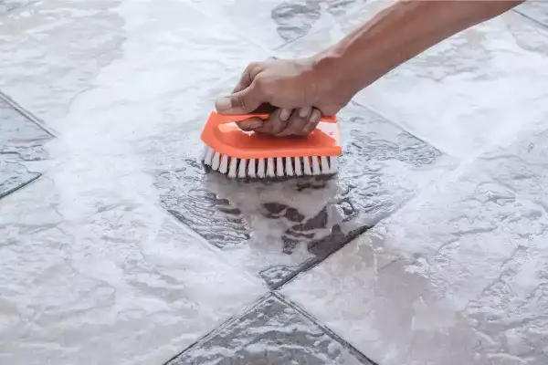 Main tenant nettoie une tache acide sur carrelage avec une brosse orange.