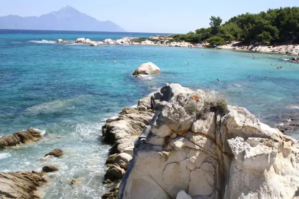 Słoneczne Chalkidiki plaże z turkusową wodą, skałami i ludźmi kąpiącymi się w morzu. W oddali widać góry.