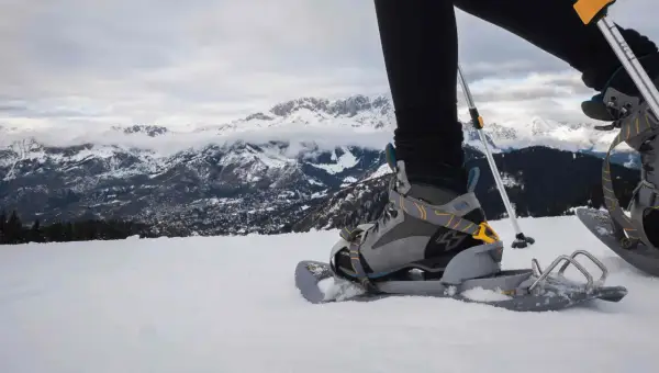 Una persona con ciaspole cammina sulla neve, con montagne innevate sullo sfondo.