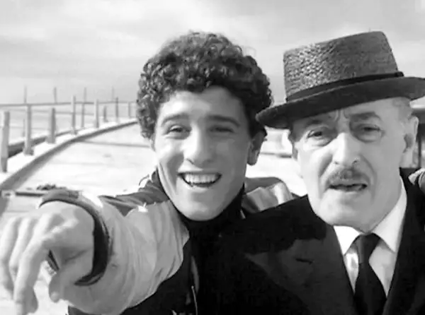 Un giovane sorridente con riccioli e un uomo anziano con cappello e baffi, un'immagine che trasmette un senso di complicità e un significato di legame generazionale.