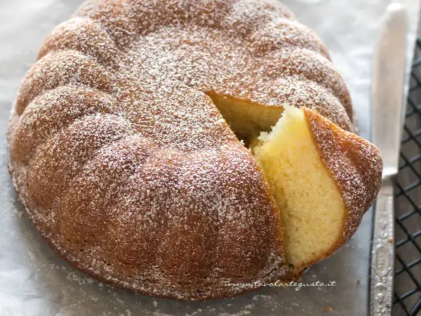 Una soffice torta allo yogurt, cosparsa di zucchero a velo, con una fetta tagliata che ne rivela l'interno dorato e profumato.